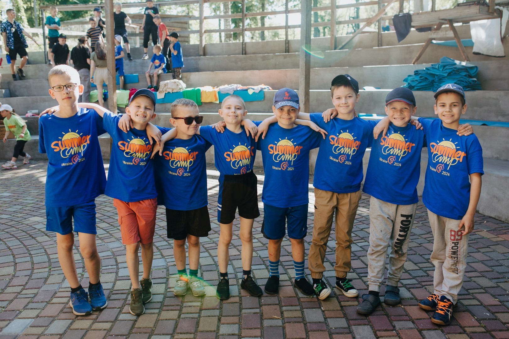 Group of young boys wearing matching camp t-shirts, smiling with arms around each other’s shoulders.
